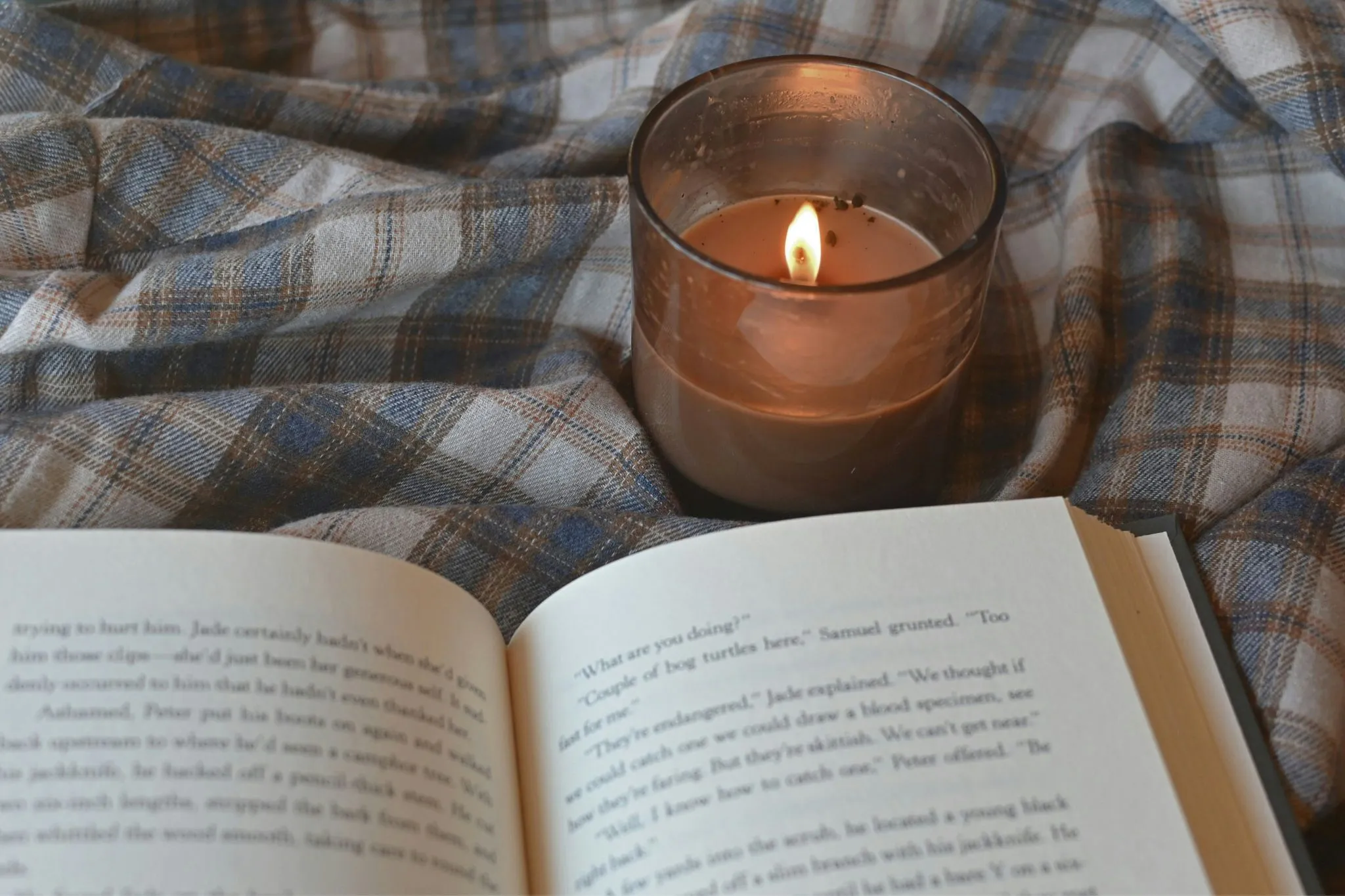 A lit candle beside an open book on a plaid blanket.
