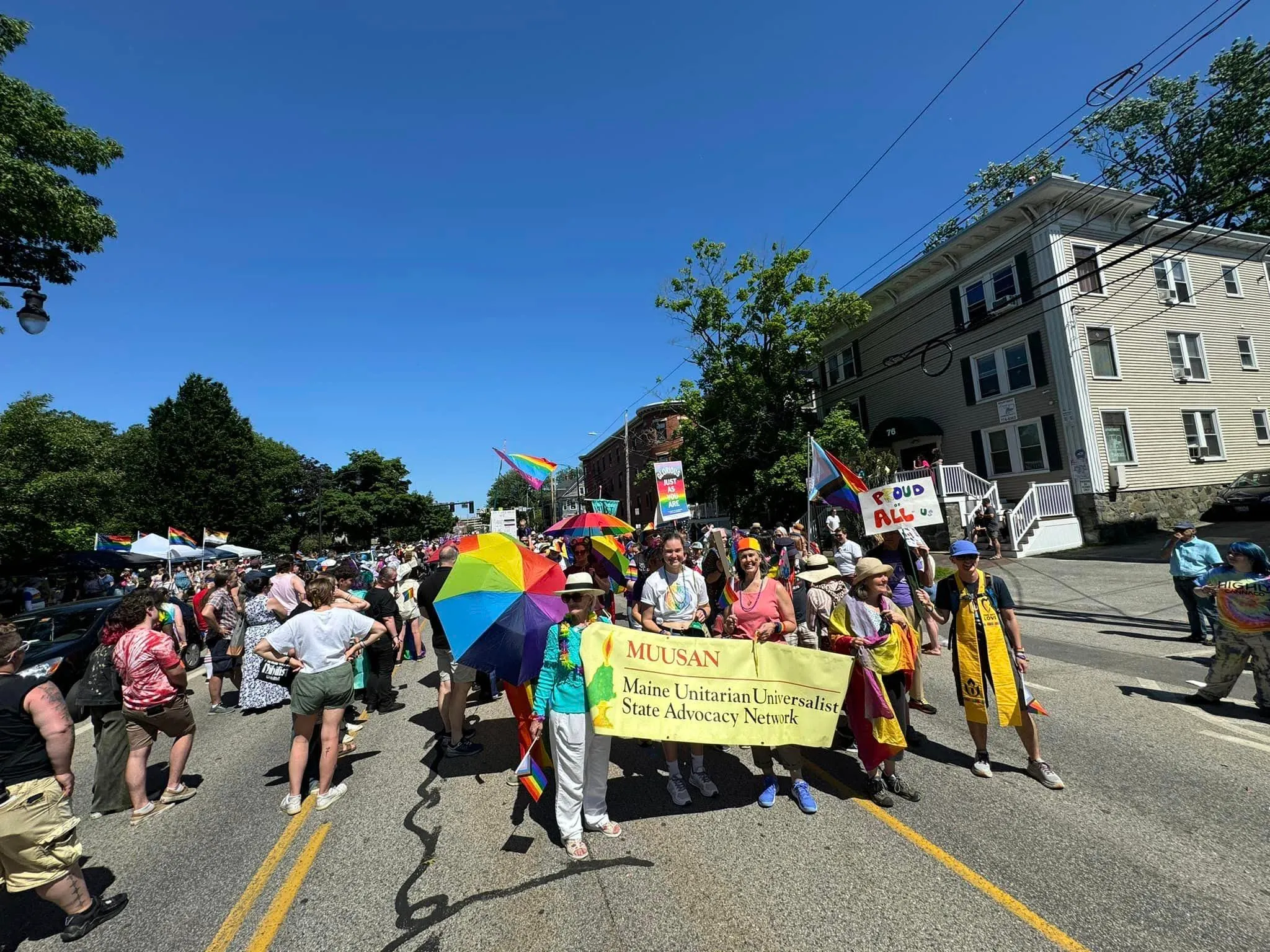 A2U2 marchers at Pride behind the yellow MUUSAN banner, with a large rainbow umbrella under a blue sky